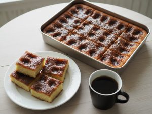 A tray of brunsviger cut into squares, its glossy dark-brown sugar and butter topping sunk into rows of dimples, with four squares on a white plate and a black mug of coffee beside it
