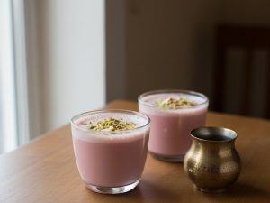 Two glasses of pale pink Kashmiri chai with a frothy top and crushed pistachios and almonds, beside a small brass pot on a wooden table